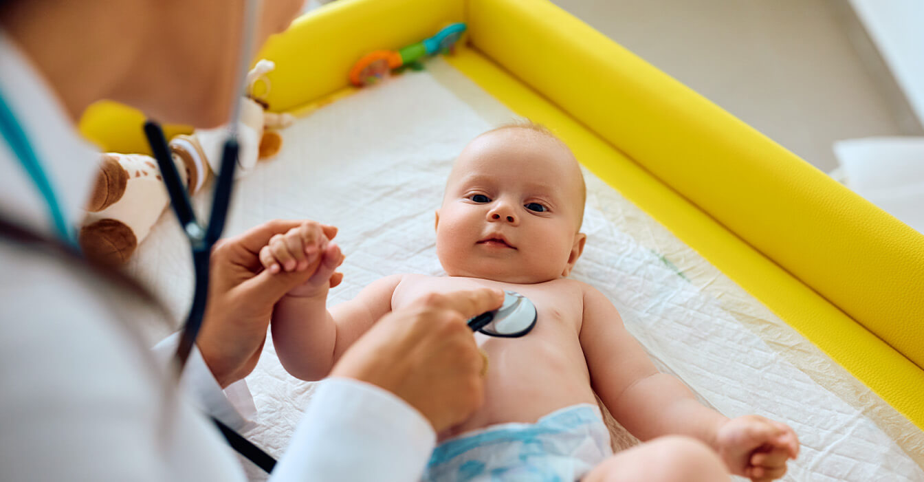 Ein Baby liegt auf einer Wickelauflage und wird von einem Arzt oder einer Ärztin, die im Vordergrund unscharf zu sehen ist, mit einem Stethoskop untersucht.