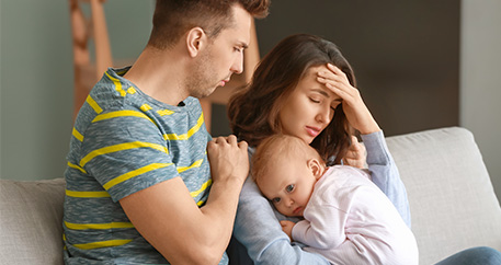 Besorgter Mann umarmt Frau, die Kopfschmerzen zu haben scheint, während sie ein kleines Kind hält, das auf ihrer Schulter ruht.