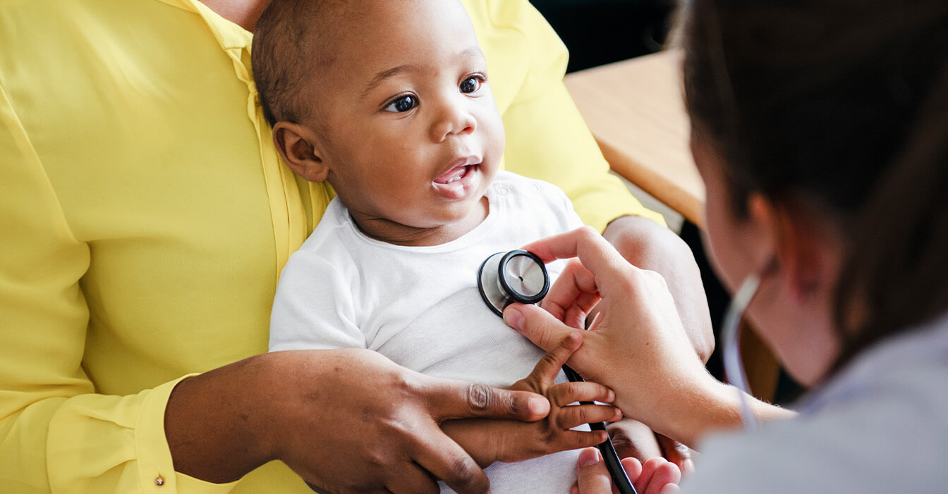 Lächelndes Baby auf dem Schoß eines Erwachsenen wird von einer Ärztin mit einem Stethoskop untersucht.
