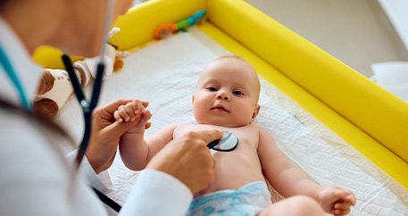  Ein Baby liegt auf einer Wickelauflage und wird von einem Arzt oder einer Ärztin mit einem Stethoskop untersucht.