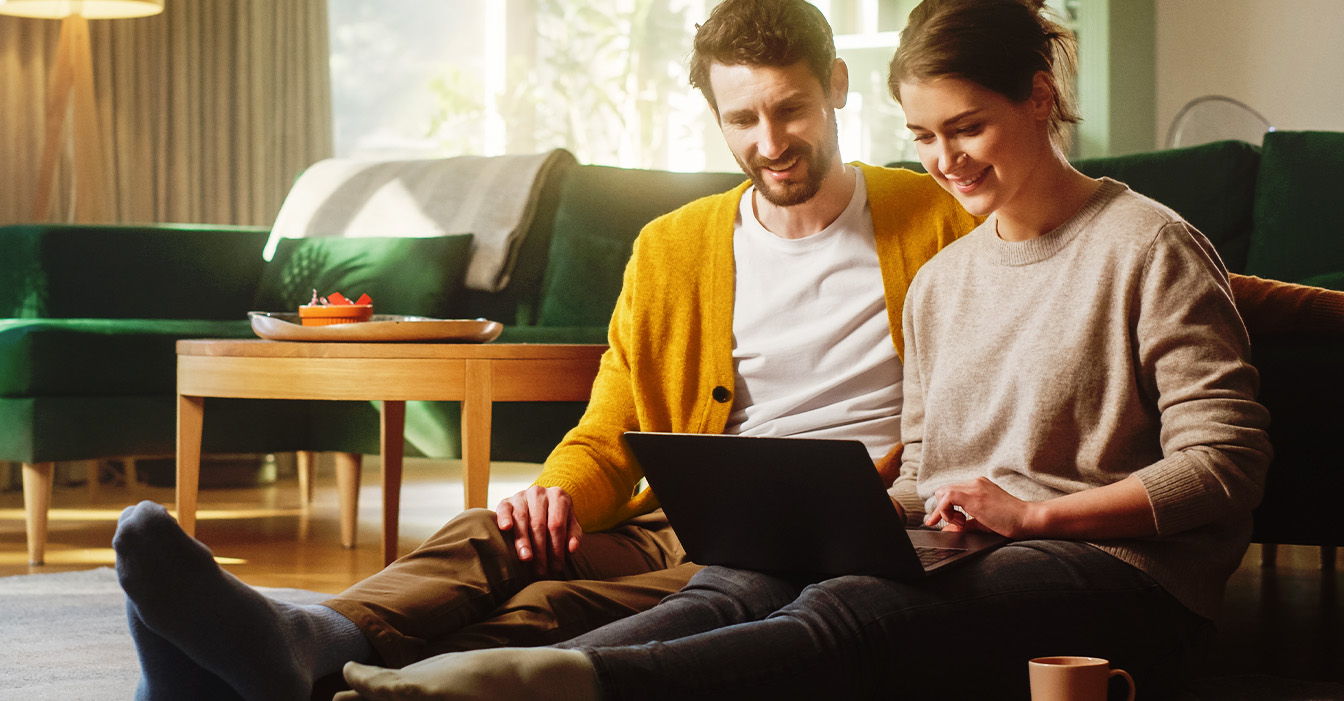  Ein Paar sitzt entspannt auf dem Boden, der Mann in einem gelben Cardigan und die Frau in einem grauen Pullover, während sie gemeinsam auf einen Laptop schauen.
