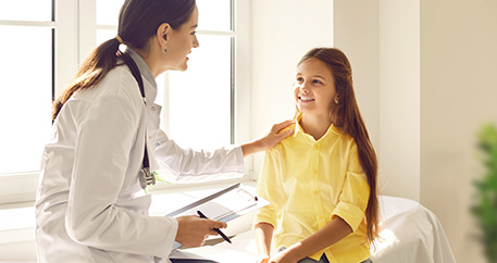  Eine freundliche Ärztin spricht mit einem lächelnden Mädchen und fasst sie dabei an der Schulter an.