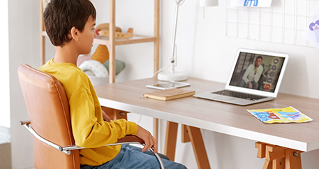 Ein Junge sitzt auf einem Stuhl an einem Schreibtisch, auf dem ein Laptop mit einem Arzt zu sehen ist.