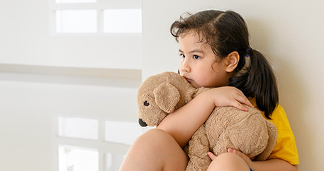 Trauriges kleines Mädchen umarmt tröstend ihren Teddybären und sitzt auf dem Boden gegen eine Wand gelehnt.