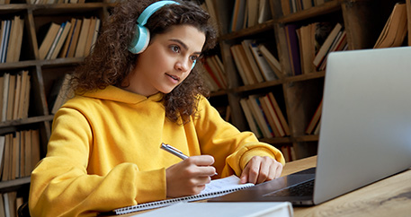 Eine junge Person mit lockigem Haar und Kopfhörern sitzt in einem gelben Pullover vor einem Laptop und schreibt in ein Notizbuch, umgeben von Bücherregalen im Hintergrund.