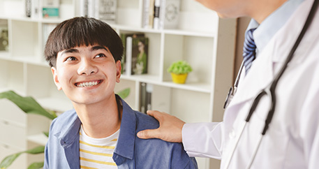  Ein lächelnder Jugendlicher schaut aufwärts zu einem Arzt, der seine Hand auf die Schulter des Jugendlichen legt in einer freundlichen Geste während eines Beratungsgesprächs.
